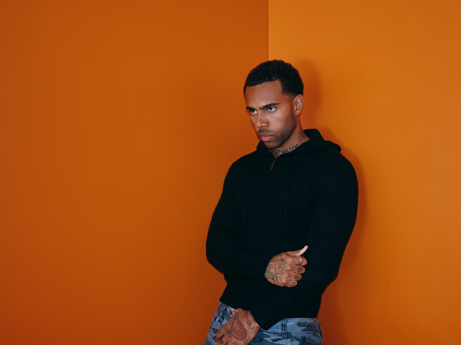 Vic Mensa in a black sweater, arms crossed, against an orange wall.