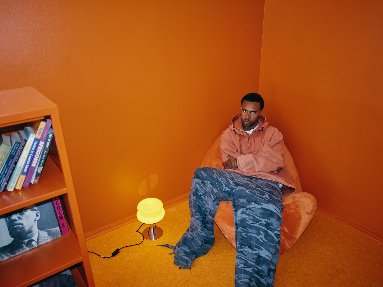 Vic Mensa reclined in an orange beanbag chair on the show's set, beside an orange bookshelf holding a Malcolm X record and a glowing yellow lamp.